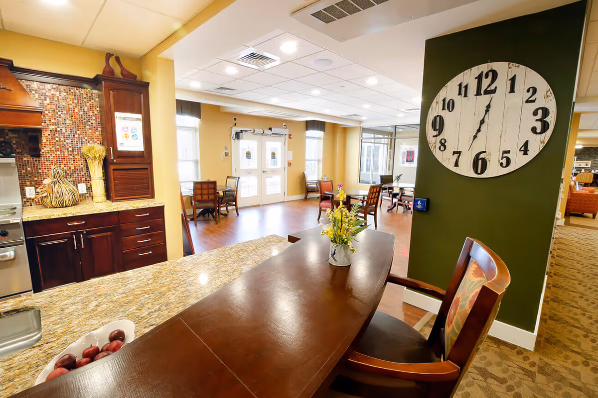 Interior view of a senior living facility showing a kitchen counter with a bowl of plums and decorative items, a wooden table with a flower vase, several chairs, a large wall clock on a green wall, and a dining area with tables and chairs near double glass doors.