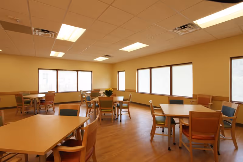 A bright dining room with multiple wooden tables and chairs arranged neatly. The room has large windows allowing natural light to fill the space, beige walls, and a wooden floor. A small plant is placed on one of the tables.