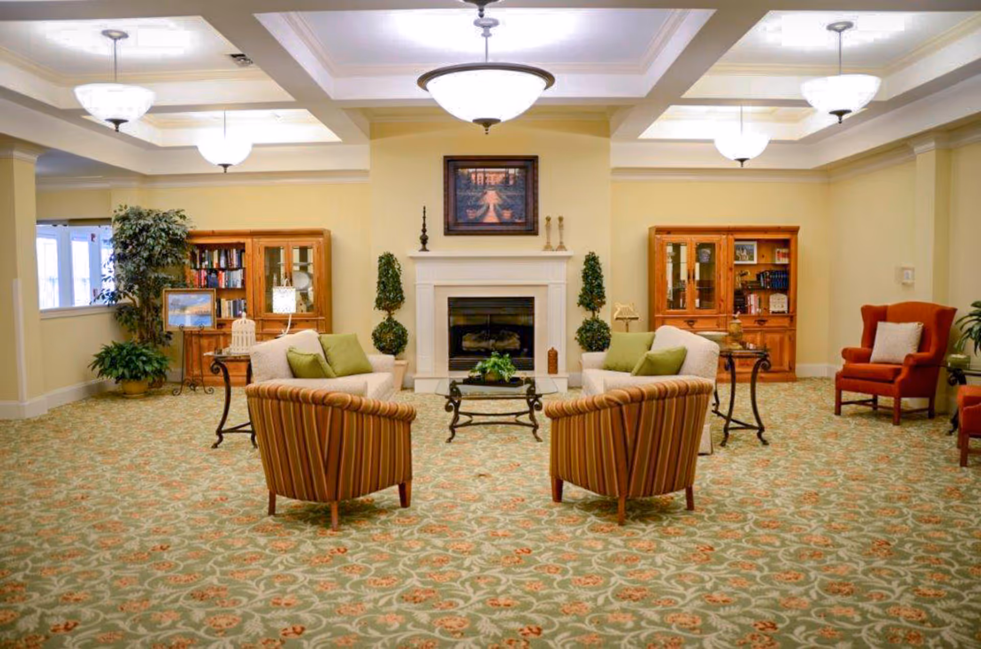 A spacious living room with patterned carpet, two striped armchairs, two beige sofas with green cushions, a glass coffee table, and a fireplace with decorative plants on either side. Wooden cabinets with books and decorative items flank the fireplace, and there are ceiling lights and a framed picture above the fireplace.