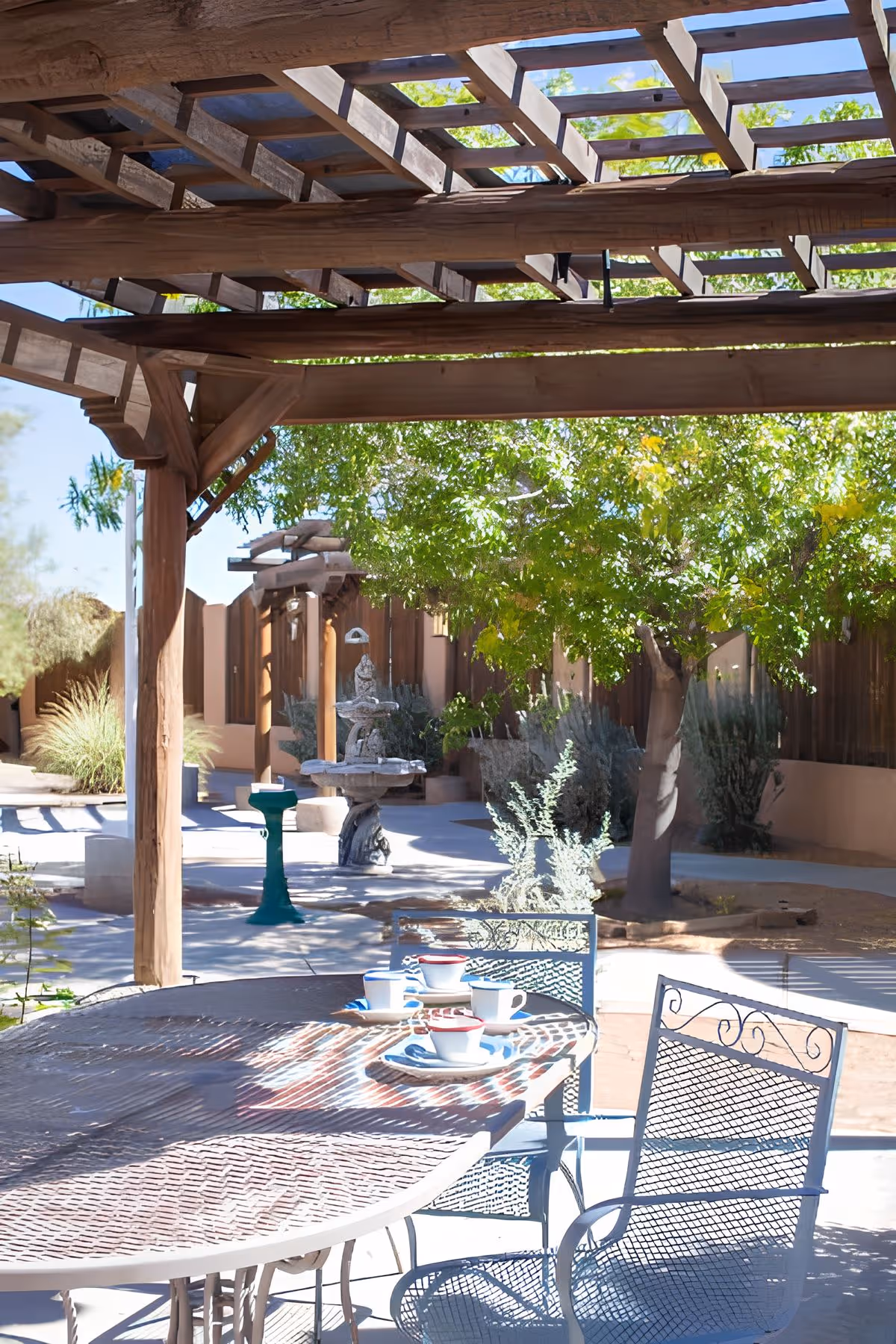 Shaded outdoor patio with a wooden pergola, a metal table and chairs set with cups, and a courtyard featuring trees and a fountain.