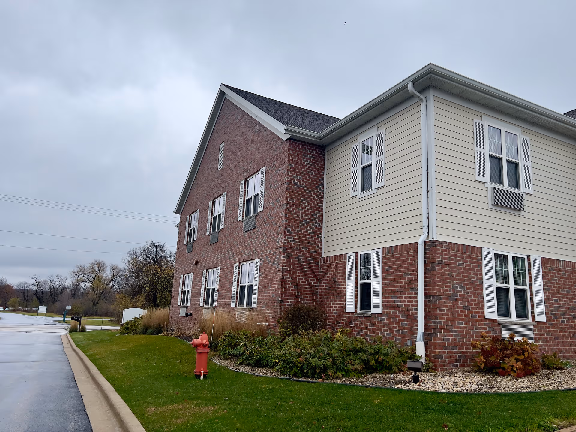Exterior view of a two-story assisted living facility building with a combination of red brick and beige siding. The building has multiple white-framed windows with white shutters. There is a green lawn with bushes and a red fire hydrant in the foreground. The sky is overcast.