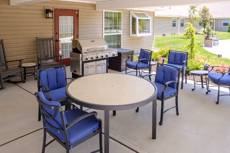Outdoor patio area with a round table surrounded by six blue cushioned chairs, two wooden rocking chairs, a stainless steel grill, and a small side table. The patio is adjacent to a beige building with a red door and windows, and there is a grassy garden area with plants and flowers nearby.