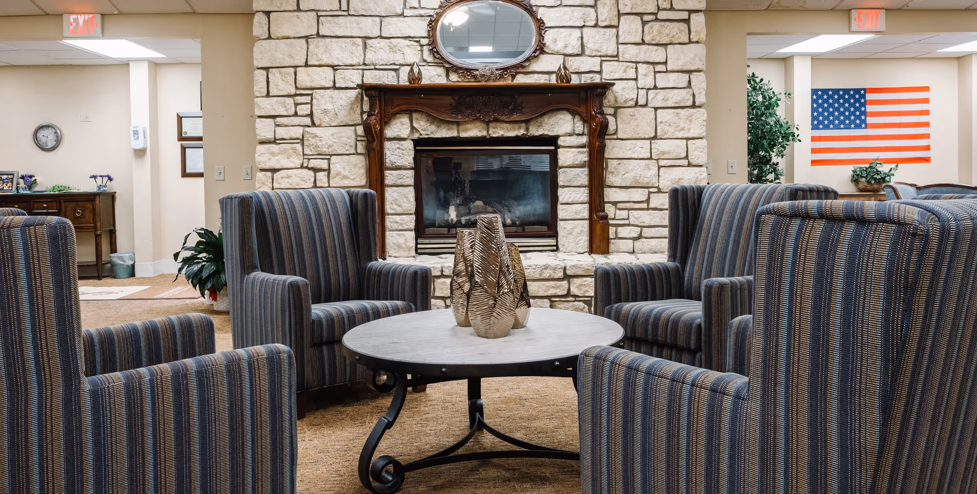 A cozy senior living facility common area with four striped armchairs arranged around a round coffee table with decorative vases. Behind the seating area is a stone fireplace with a wooden mantel and an oval mirror above it. The room has beige walls, carpeted floor, an American flag on the wall, and some plants and furniture in the background.