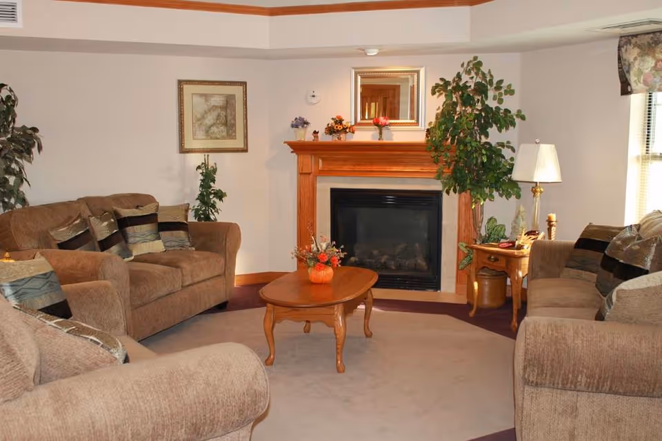 A cozy living room with three brown upholstered sofas arranged around a wooden coffee table with a small floral centerpiece. The room features a fireplace with a wooden mantel decorated with small flower pots and a mirror above it. There are two large potted plants on either side of the fireplace, a framed picture on the wall, and a table lamp on a side table near a window with blinds and a floral valance.