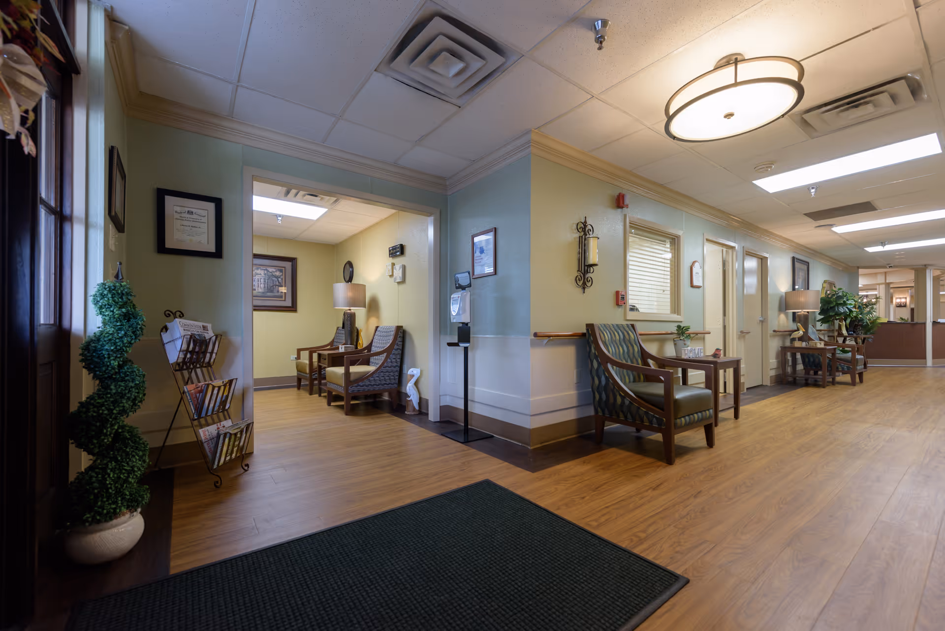 A well-lit hallway in a senior living facility with wooden flooring and light green walls. The hallway features several chairs with patterned cushions, small tables with lamps and plants, framed pictures on the walls, and a magazine rack near the entrance. Ceiling lights illuminate the space, and there is a dark green floor mat near the door.