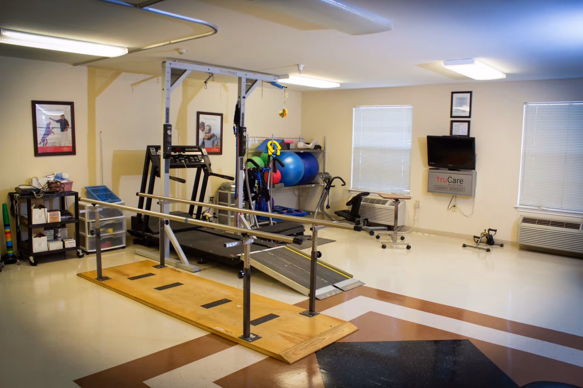 A physical therapy or rehabilitation room with parallel bars on a wooden platform, a treadmill with a ramp, exercise balls on a metal rack, and various exercise equipment. The room has two windows with blinds, two air conditioning units, framed pictures on the walls, and a wall-mounted TV with a truCare therapy services sign beneath it.