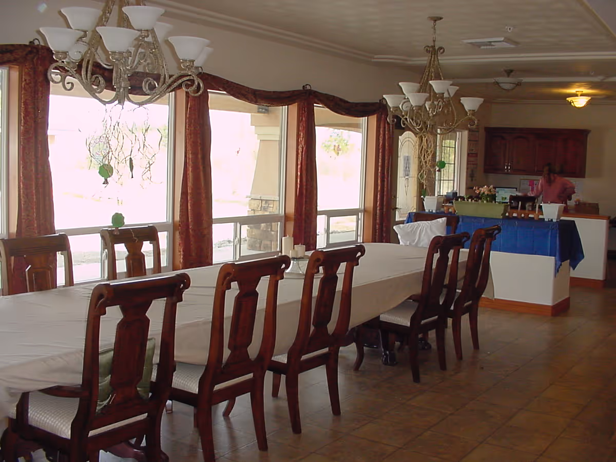 A long dining table covered with a white tablecloth surrounded by wooden chairs in a room with large windows and red curtains. Two chandeliers hang from the ceiling above the table. In the background, there is a kitchen area with wooden cabinets and a person standing near the counter.