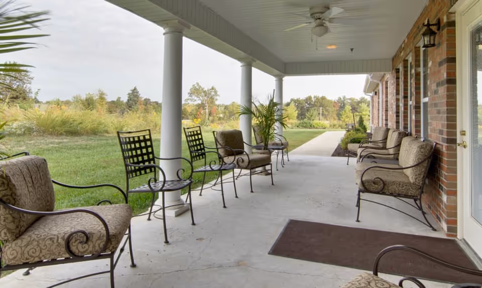 Covered outdoor patio with cushioned metal chairs along a brick building overlooking a grassy landscape.