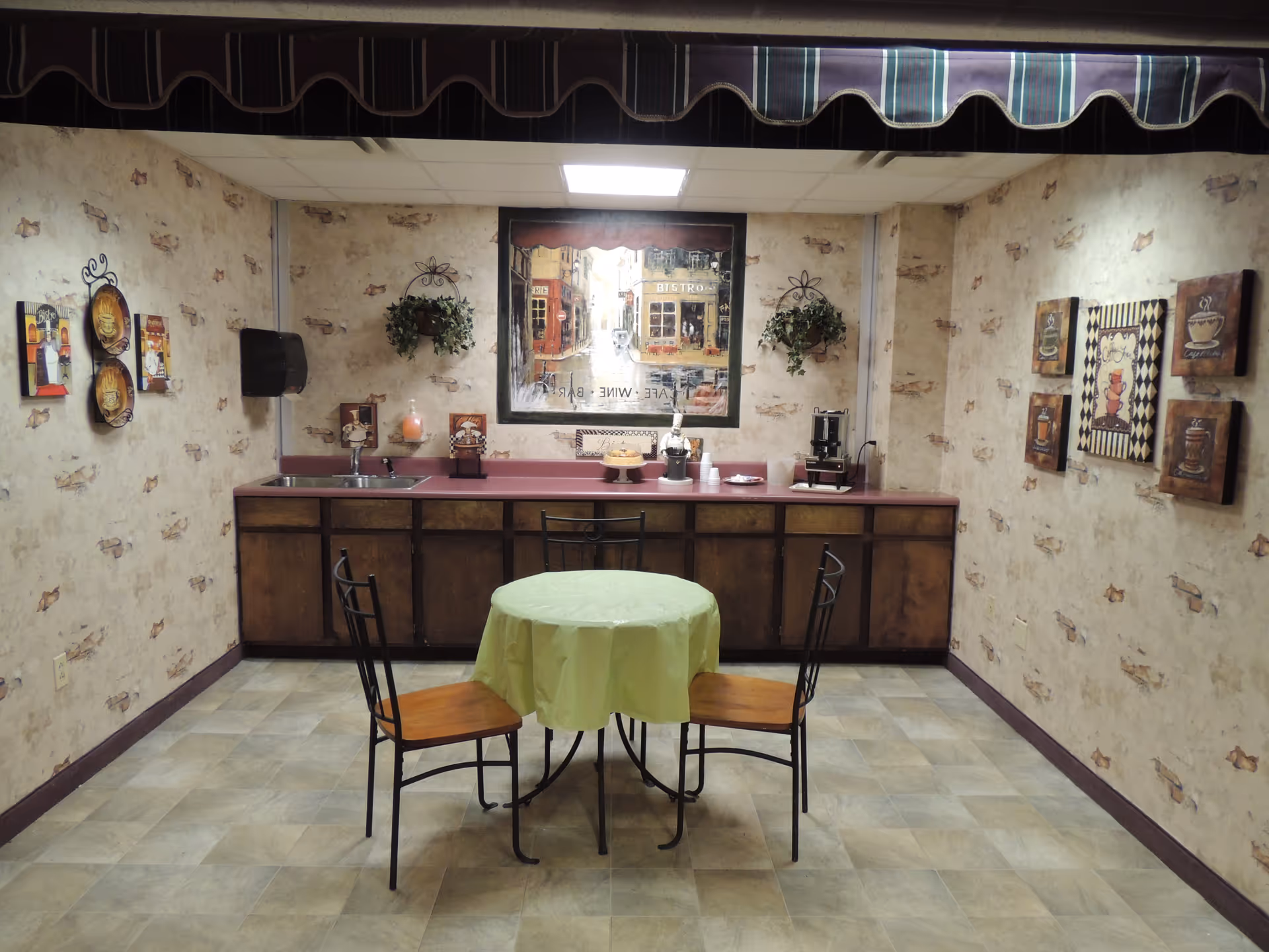 A small dining area with a round table covered with a green tablecloth and two wooden chairs with black metal frames. The room has beige wallpaper with a subtle pattern and several framed pictures on the walls. Along the back wall is a countertop with a sink, coffee maker, and various decorative items. Two hanging plants are mounted on the wall above the counter, and a large framed picture depicting a street scene is centered on the back wall.