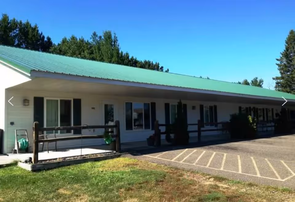 Exterior view of a single-story building with a green metal roof and white siding. The building has multiple windows and a covered porch area with wooden railings. There is a parking area with marked spaces in front and some trees in the background under a clear blue sky.