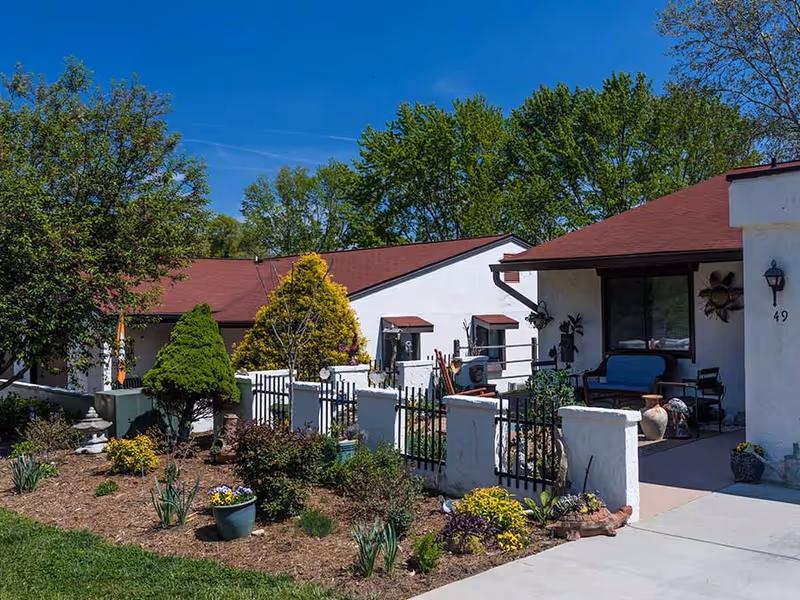 Front exterior of a single-story retirement community building with a fenced garden, patio seating, and a red roof under a clear blue sky.