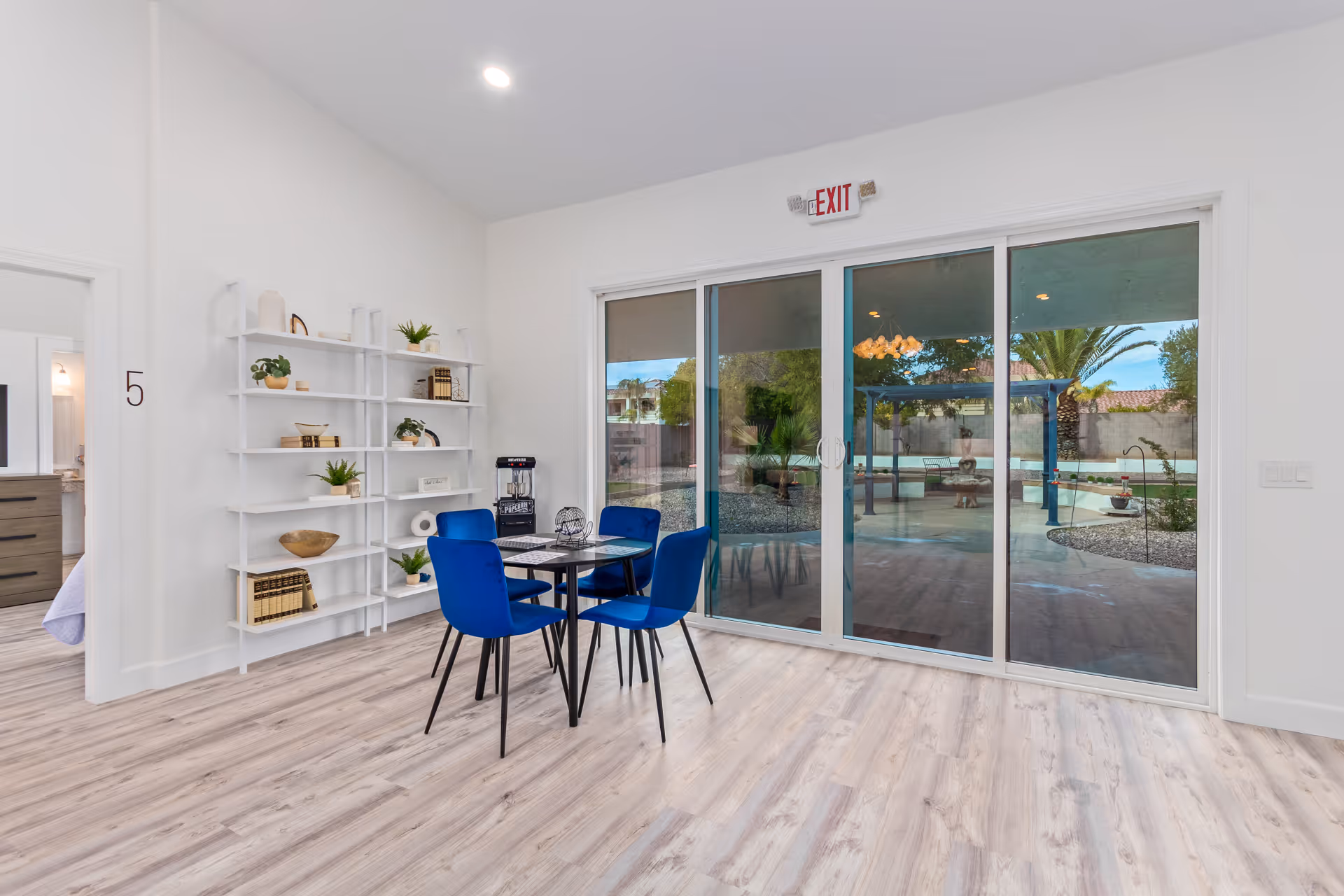 A bright interior room with light wood flooring, white walls, and a high ceiling. There is a small black dining table with four blue chairs in the center. To the left, a white shelving unit holds decorative plants, books, and bowls. Large sliding glass doors lead to an outdoor area with palm trees and a covered patio. An exit sign is above the doors.