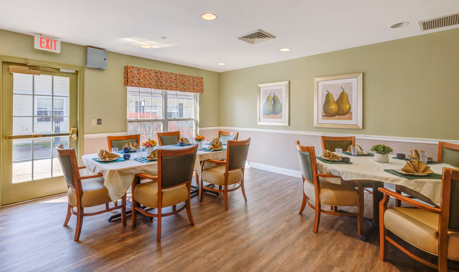 A bright dining room with two tables set for meals, each with beige tablecloths, folded napkins, and small flower arrangements. The room has light green walls with two framed pictures of pears, a window with a patterned valance, and a door with an exit sign above it. The floor is wood, and the chairs have wooden frames with beige cushions.