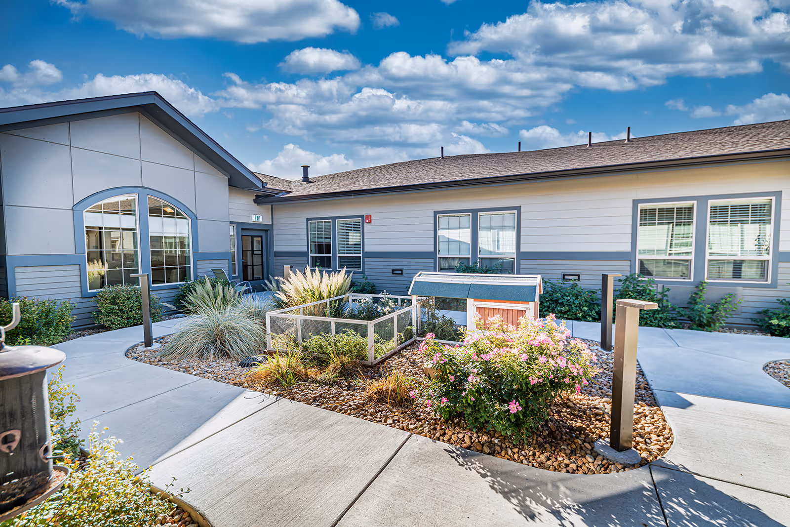 Outdoor courtyard area of a senior living facility with a paved walkway, landscaped garden beds containing ornamental grasses and flowering plants, and a small fenced garden structure. The building exterior is visible with large windows and a door under a blue sky with scattered clouds.
