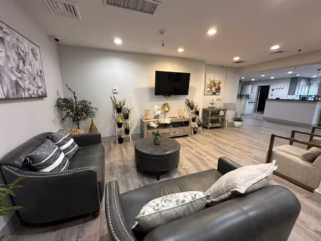 A modern living room area in a senior living facility with two black leather sofas adorned with pillows, a round black ottoman in the center, and a wall-mounted flat-screen TV above a wooden console table decorated with various ornaments and plants. The room has wood flooring, recessed ceiling lights, and neutral-colored walls with framed artwork. In the background, there is an open view into a kitchen area and additional seating with armchairs.
