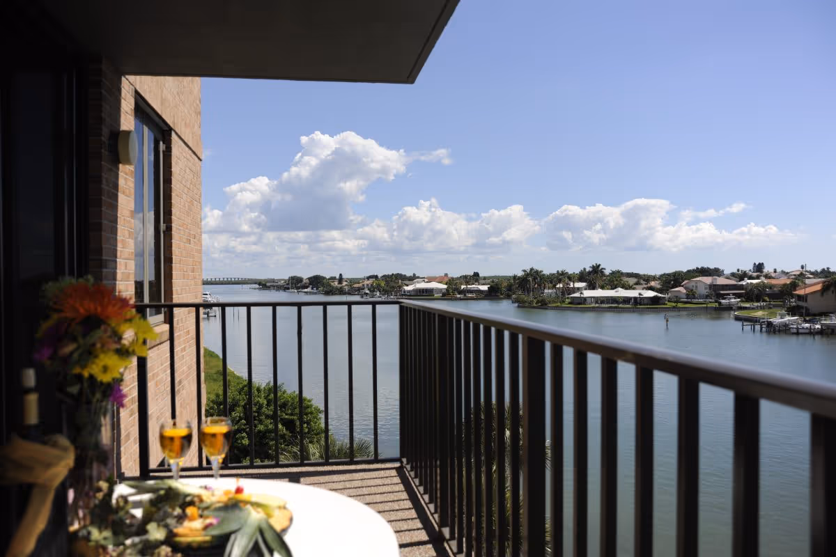 View from a balcony overlooking a calm river with houses and palm trees on the opposite shore under a blue sky with scattered clouds. The balcony has a black metal railing, and a table with two glasses of a golden beverage and a flower arrangement is partially visible.