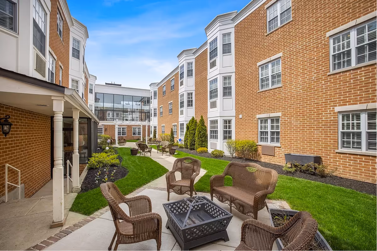 Outdoor courtyard area of a senior living facility with brick buildings on either side, green grass, landscaped plants, and several wicker chairs and benches arranged around a fire pit table on a paved patio.