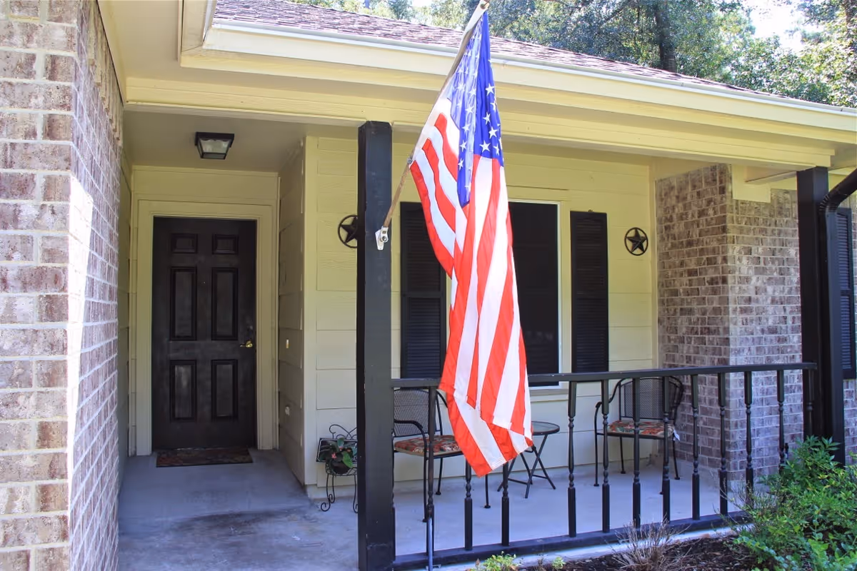 Front porch of a cottage with an American flag, a dark front door, and a small seating area with chairs.