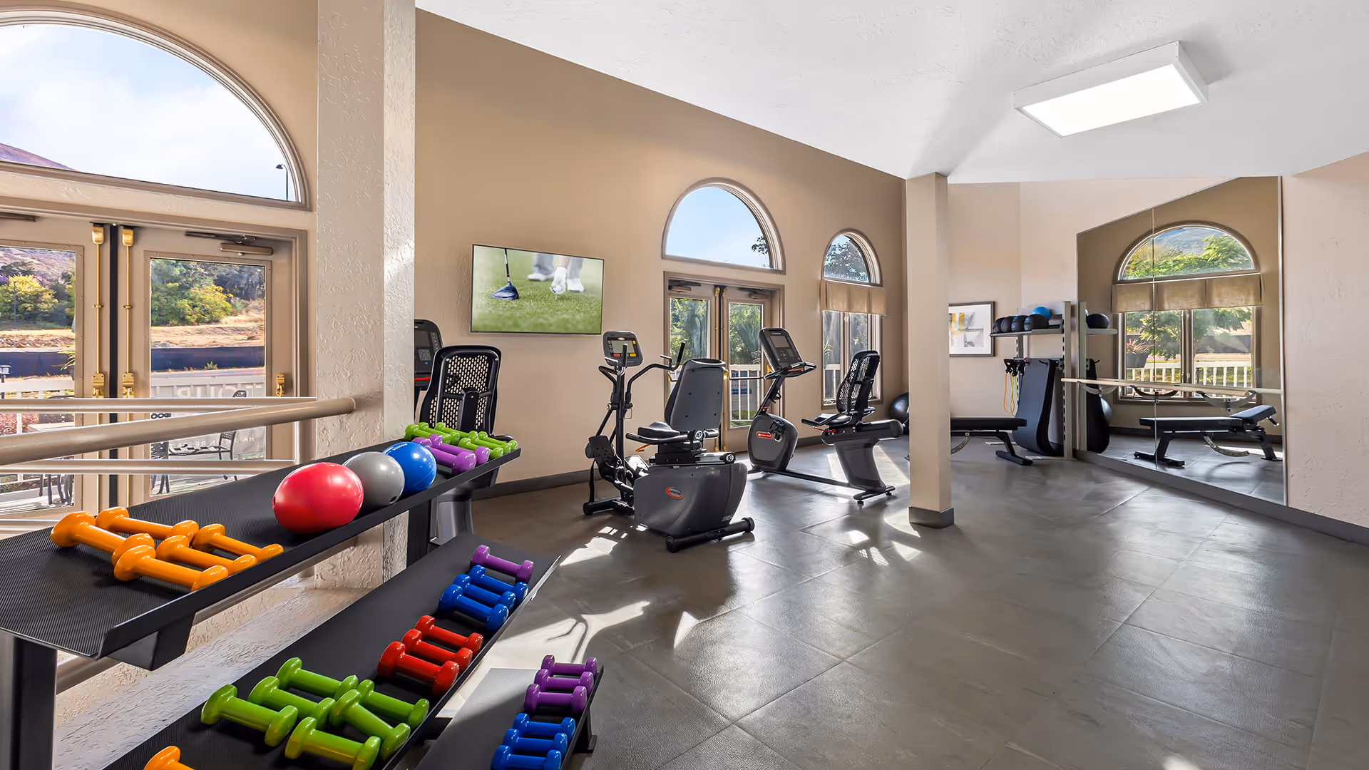 A bright fitness room with large arched windows letting in natural light. The room contains various exercise equipment including stationary bikes, a weight bench, and racks of colorful dumbbells and exercise balls. A TV mounted on the wall displays a golf scene. The floor is tiled and there is a large mirror on one wall.