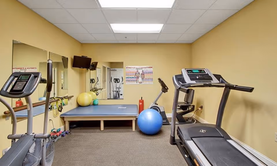 A small exercise room with a treadmill, stationary bike, elliptical machine, exercise balls, dumbbells, and a raised platform. The walls are painted yellow and there are mirrors and a TV mounted on the wall.