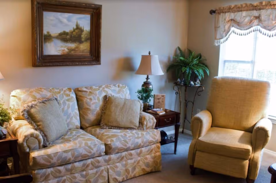A cozy living room area with a patterned loveseat featuring two beige cushions, a beige armchair, a wooden side table with a lamp and small plants, a framed landscape painting on the wall, and a window with a decorative valance.