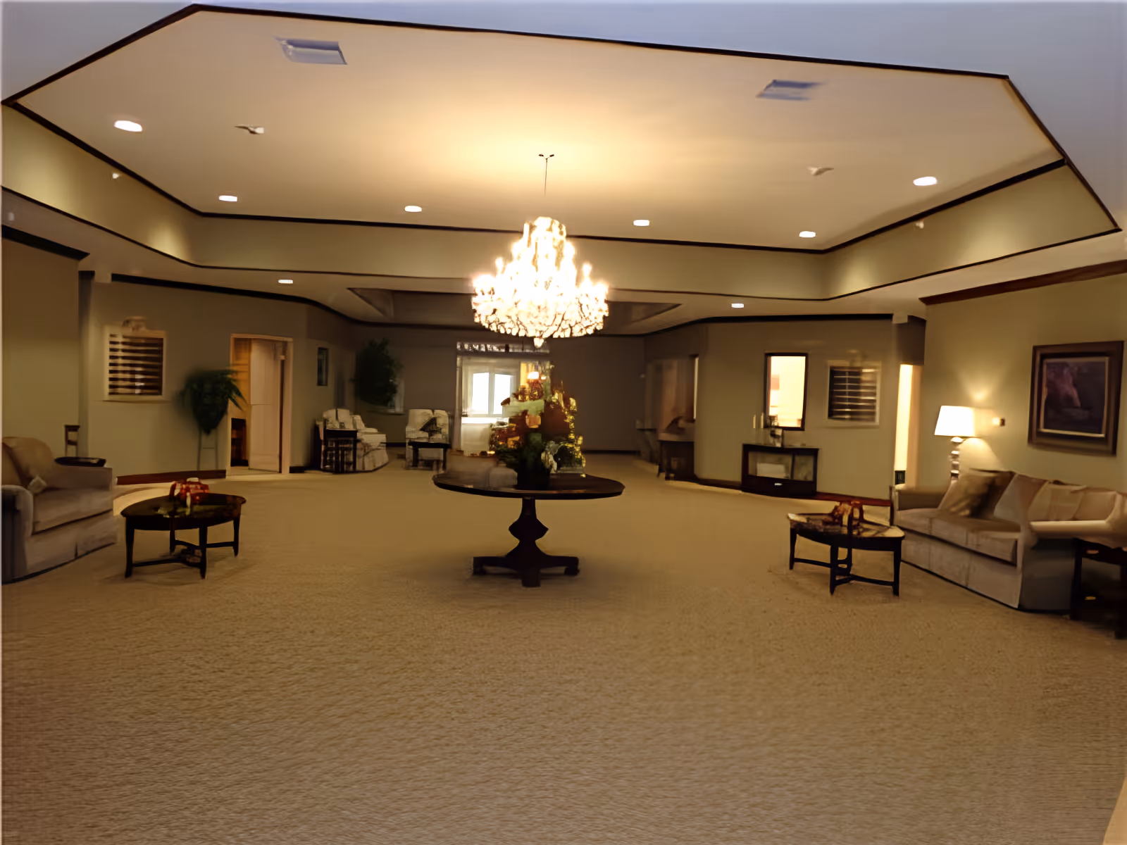 A spacious senior living community lounge area with beige carpeting, neutral-colored walls, and a large ornate chandelier hanging from the ceiling. The room features multiple seating areas with sofas and armchairs, small tables with floral arrangements, framed artwork on the walls, and soft lighting from lamps.