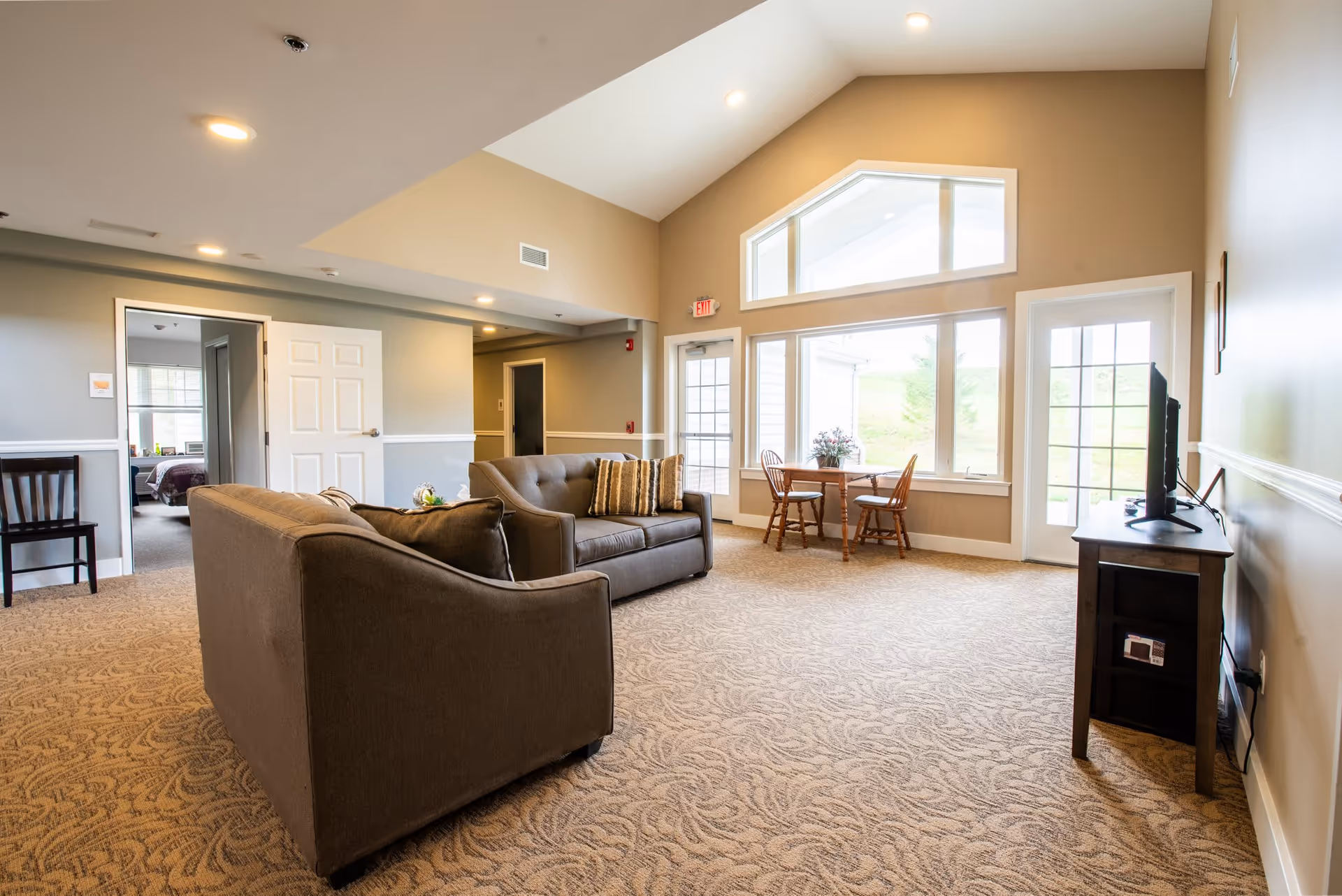 A spacious living room with beige walls and carpeted floor featuring two gray sofas with cushions, a small wooden table with two chairs near large windows, and a TV on a stand. There is a door leading outside and an open doorway showing a bedroom in the background.