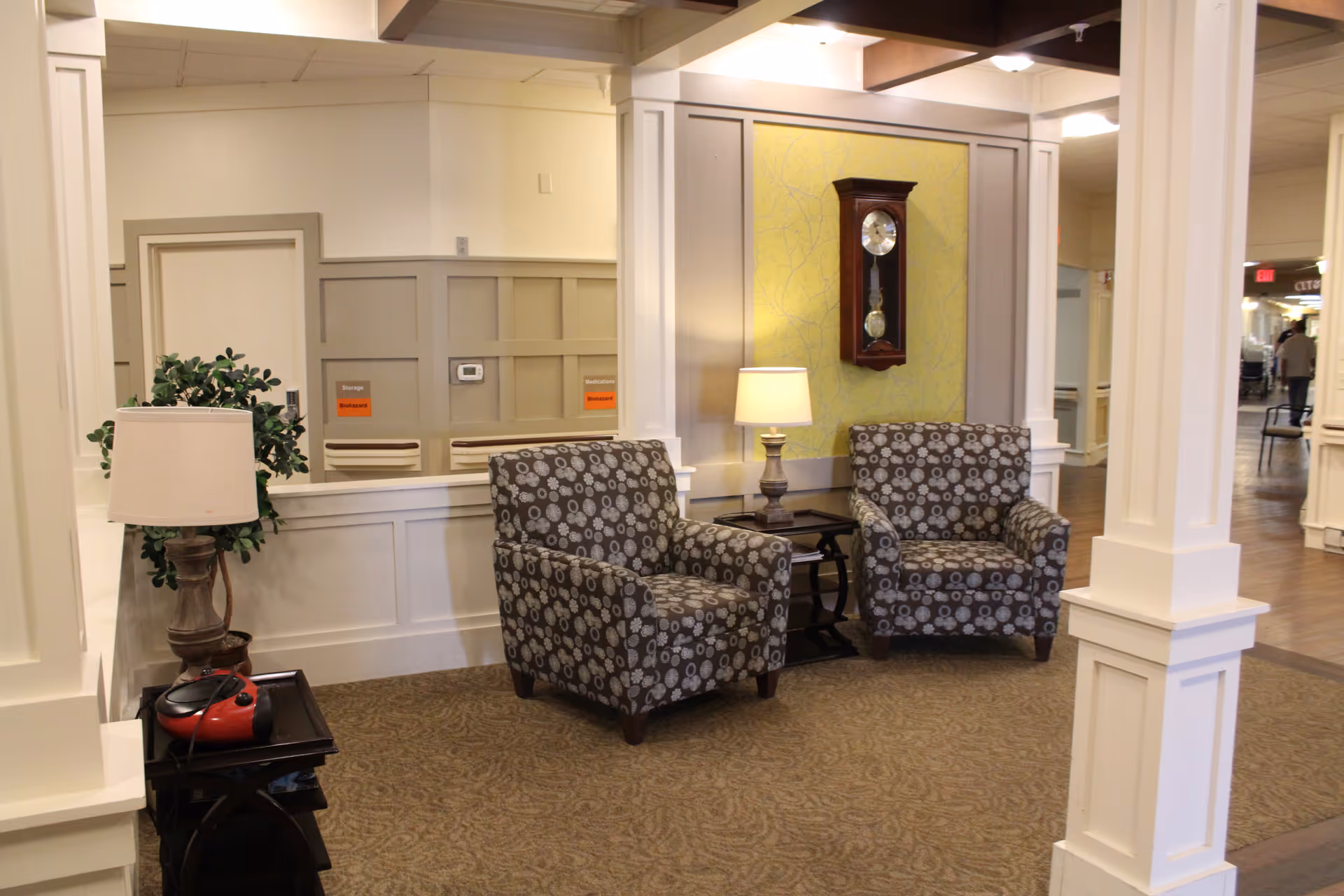 A cozy seating area in a senior living facility with two patterned armchairs, a small table with a lamp between them, a wall clock mounted on a yellow accent wall, and a side table with a lamp and a red device on it. The space has beige walls with white trim and carpeted flooring.
