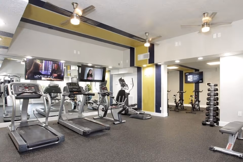 A modern fitness room with several treadmills, stationary bikes, and elliptical machines. There are two wall-mounted televisions above the treadmills, a rack of dumbbells, and a bench. The room has ceiling fans, recessed lighting, and a large mirror along one wall.