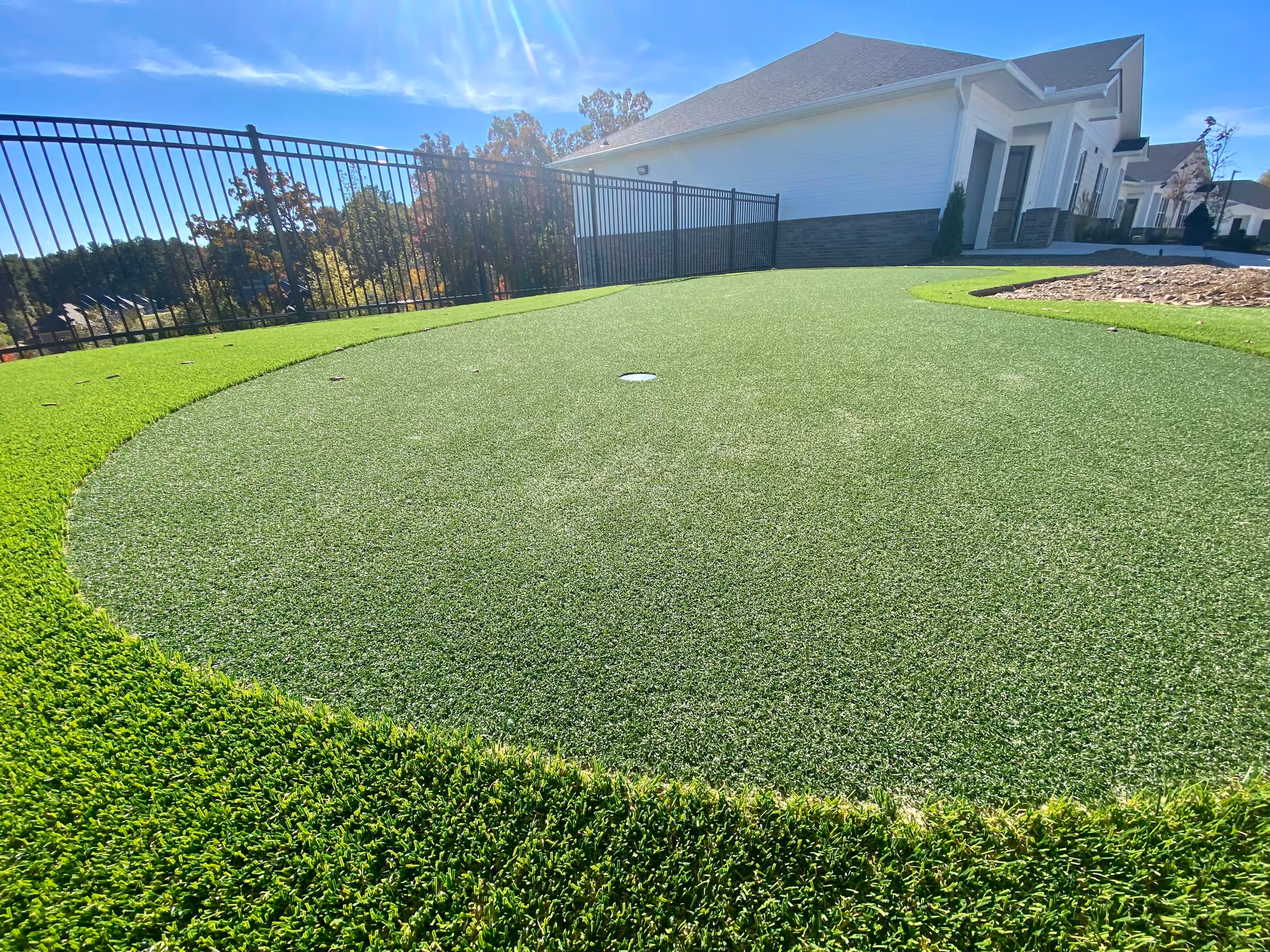 Outdoor view of a putting green area with artificial turf, surrounded by a black metal fence. In the background, there is a white building with a stone base and a clear blue sky with sunlight.