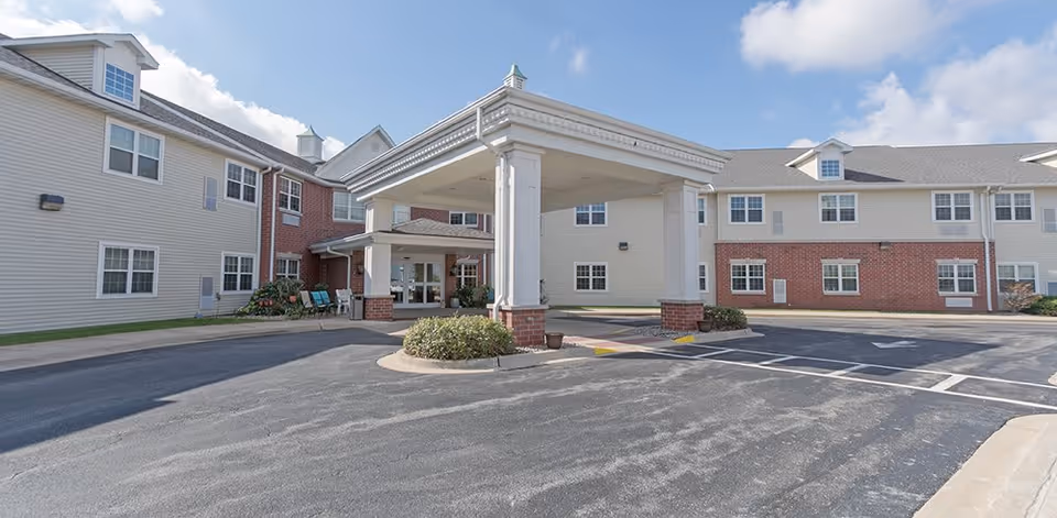 Exterior view of Heritage Woods of Dwight senior living facility showing a two-story building with beige siding and red brick accents, a covered entrance with white columns, and a paved driveway under a partly cloudy sky.