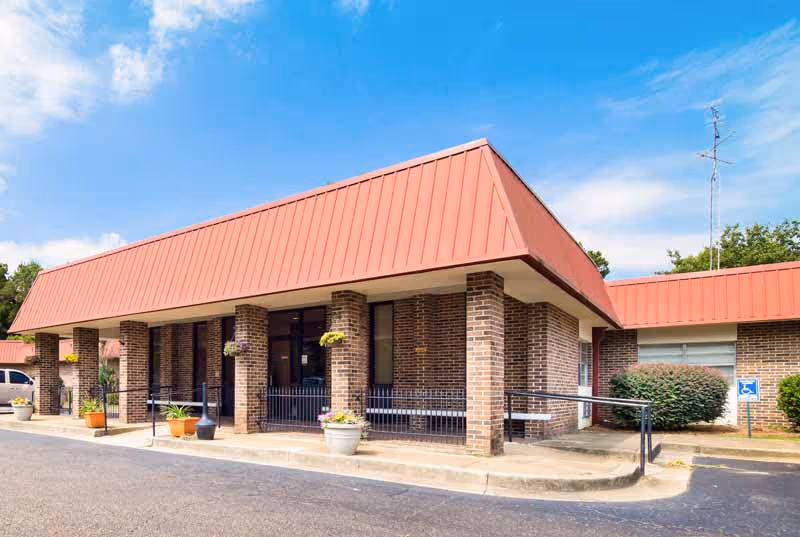 Exterior view of a single-story brick building with a red sloped roof and multiple columns supporting a covered entrance. There are potted plants and hanging flower baskets near the entrance, a wheelchair accessible ramp, and a parking area in front under a partly cloudy sky.