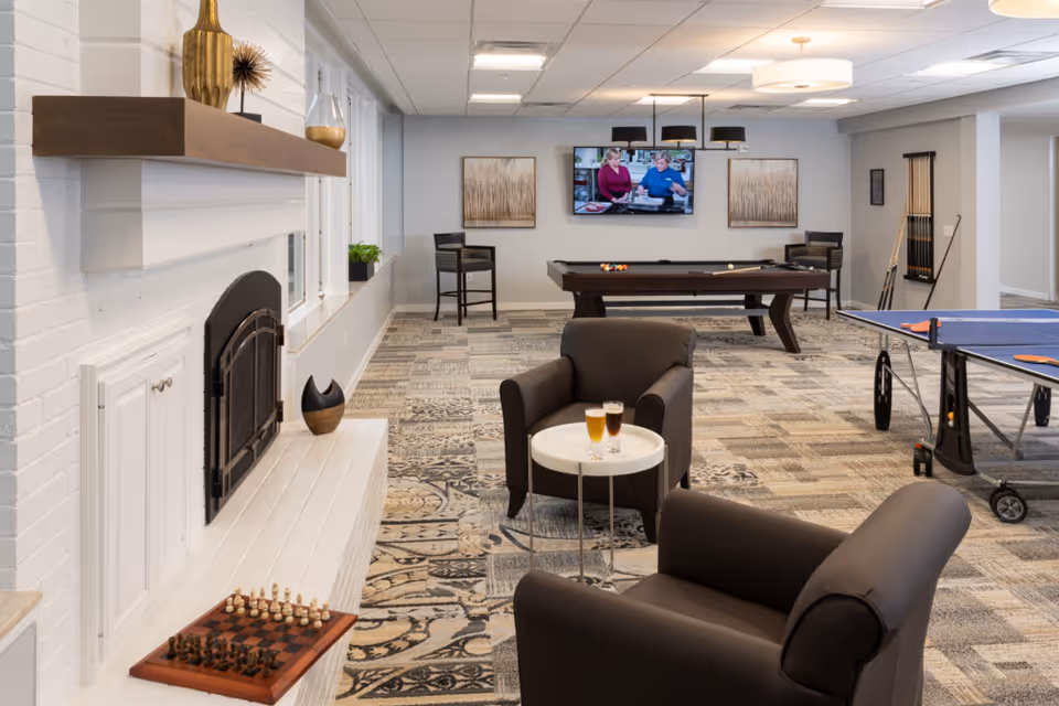 A spacious recreational room with a white brick fireplace on the left, two dark armchairs with a small round table holding two glasses of beer, a chessboard on the fireplace hearth, a pool table with balls and cues, a ping pong table with paddles and balls, and a wall-mounted TV showing two people talking. The room has patterned carpet flooring, modern ceiling lights, and framed artwork on the walls.