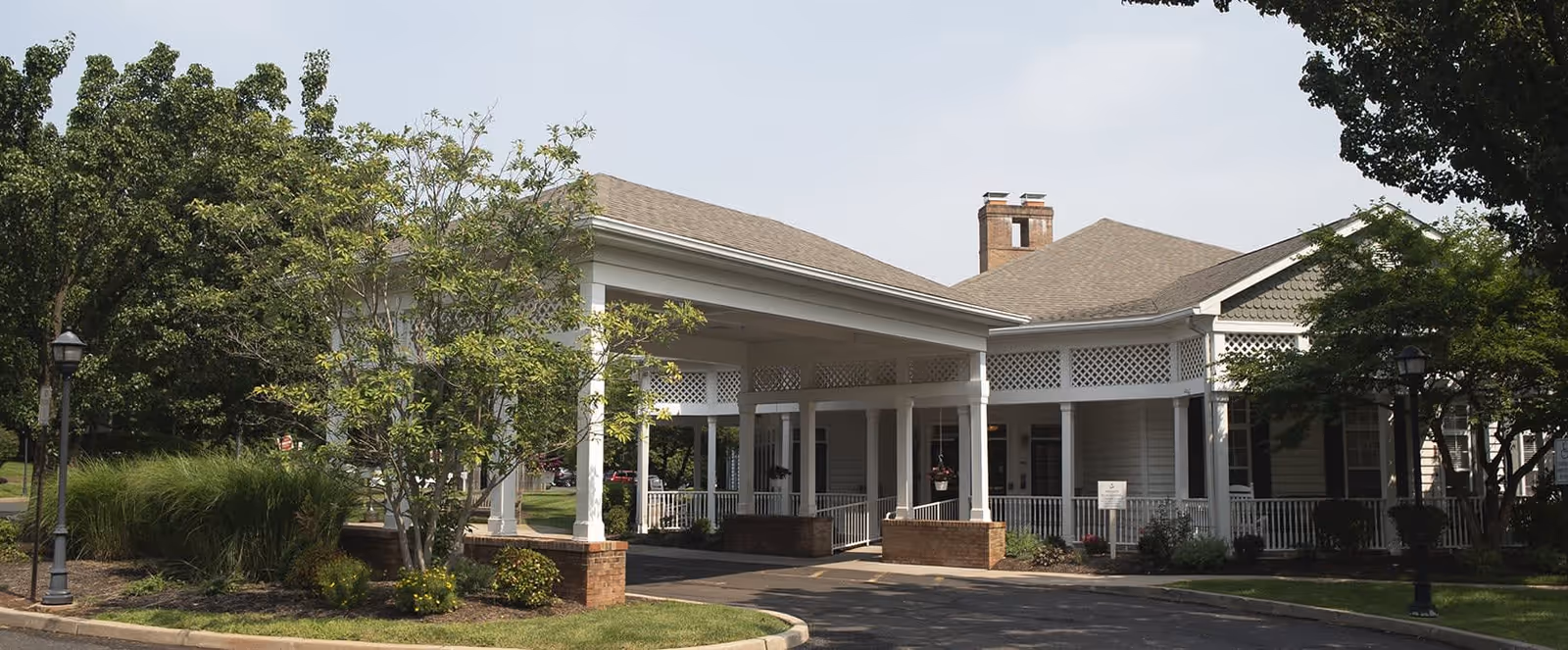 Exterior view of a single-story building with a covered entrance supported by white columns, surrounded by trees and landscaping under a clear sky.