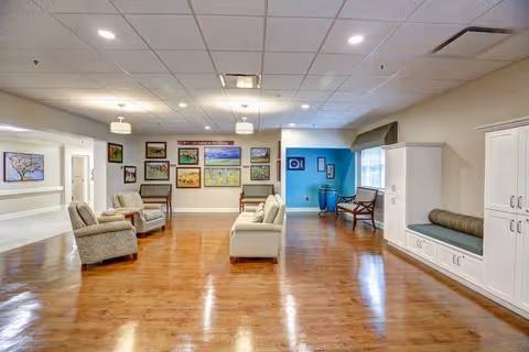 A spacious, well-lit common area with wooden flooring and a drop ceiling with recessed lighting. The room features comfortable seating including armchairs and a sofa arranged around a small table. The walls are decorated with framed artwork and paintings. There is a built-in bench with cushions and storage cabinets along one wall, and a window with a valance letting in natural light. The overall atmosphere is warm and inviting.