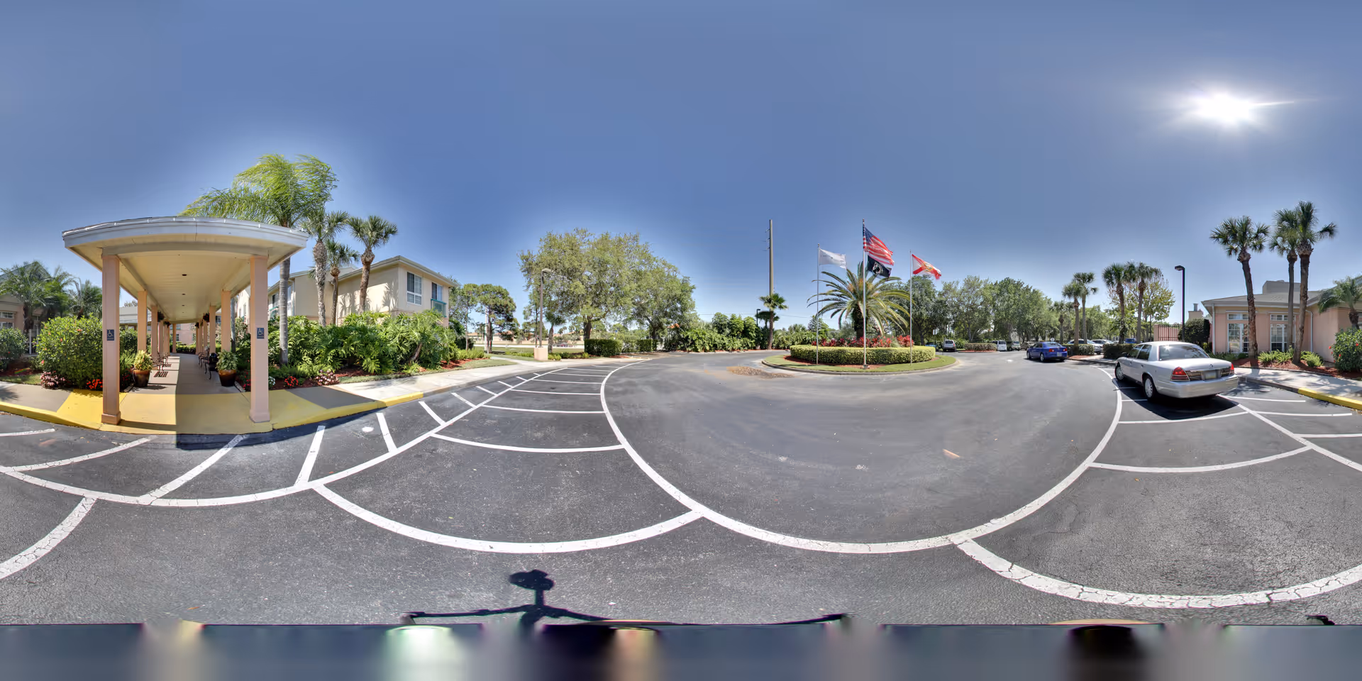 Wide panoramic view of the parking lot and entrance area of The Gardens of Port St. Lucie senior living facility on a sunny day, showing a covered walkway, several palm trees, landscaped greenery, flags on flagpoles, and parked cars.