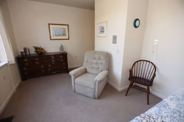 A simple room with a beige armchair and a wooden chair against white walls. There is a wooden dresser with decorative items and framed pictures hanging on the walls. A bed with a floral bedspread is partially visible on the right side.