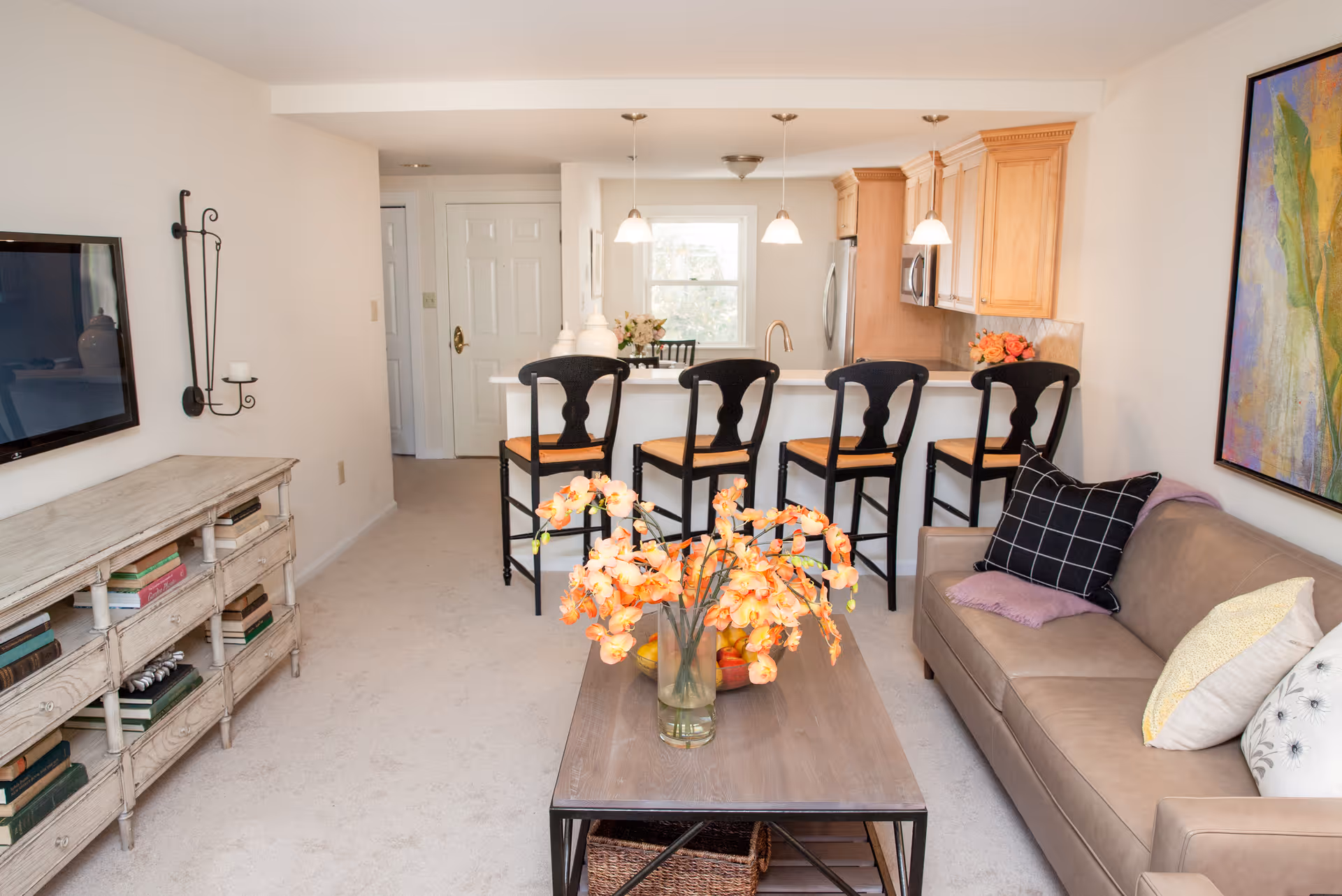 Open-plan living room and kitchen with a sofa, coffee table holding a vase of flowers, and four bar stools at a breakfast bar.