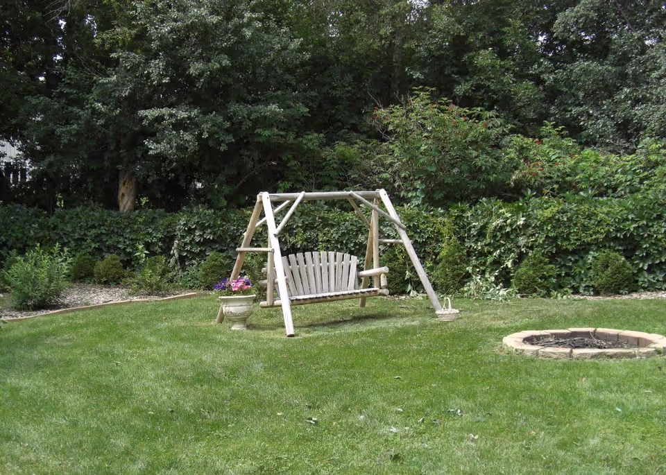 A wooden garden swing set on a green lawn with a flower pot beside it and a circular stone fire pit on the right. The background is filled with dense green bushes and trees.