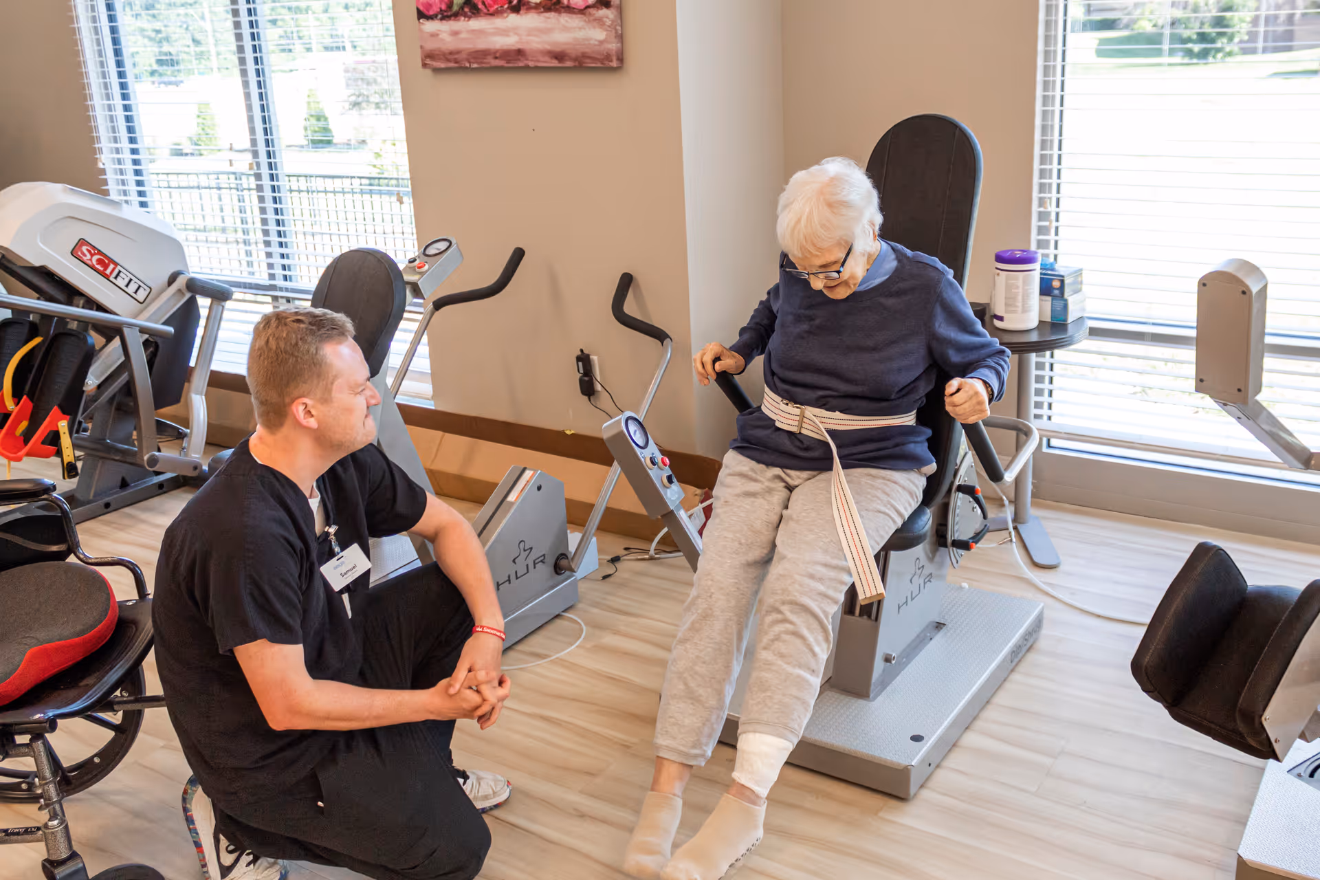 An elderly woman seated on a rehabilitation exercise machine with a strap around her waist, assisted by a male healthcare worker kneeling beside her in a bright room with exercise equipment and large windows.
