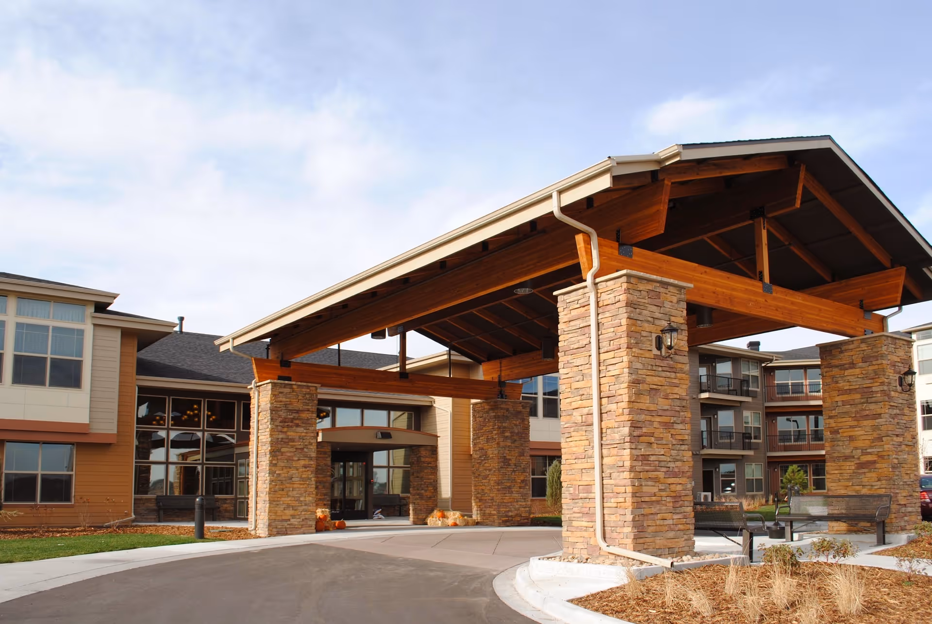 Covered entrance with a wooden-beamed porte-cochere supported by stone pillars at the front of a senior living building.