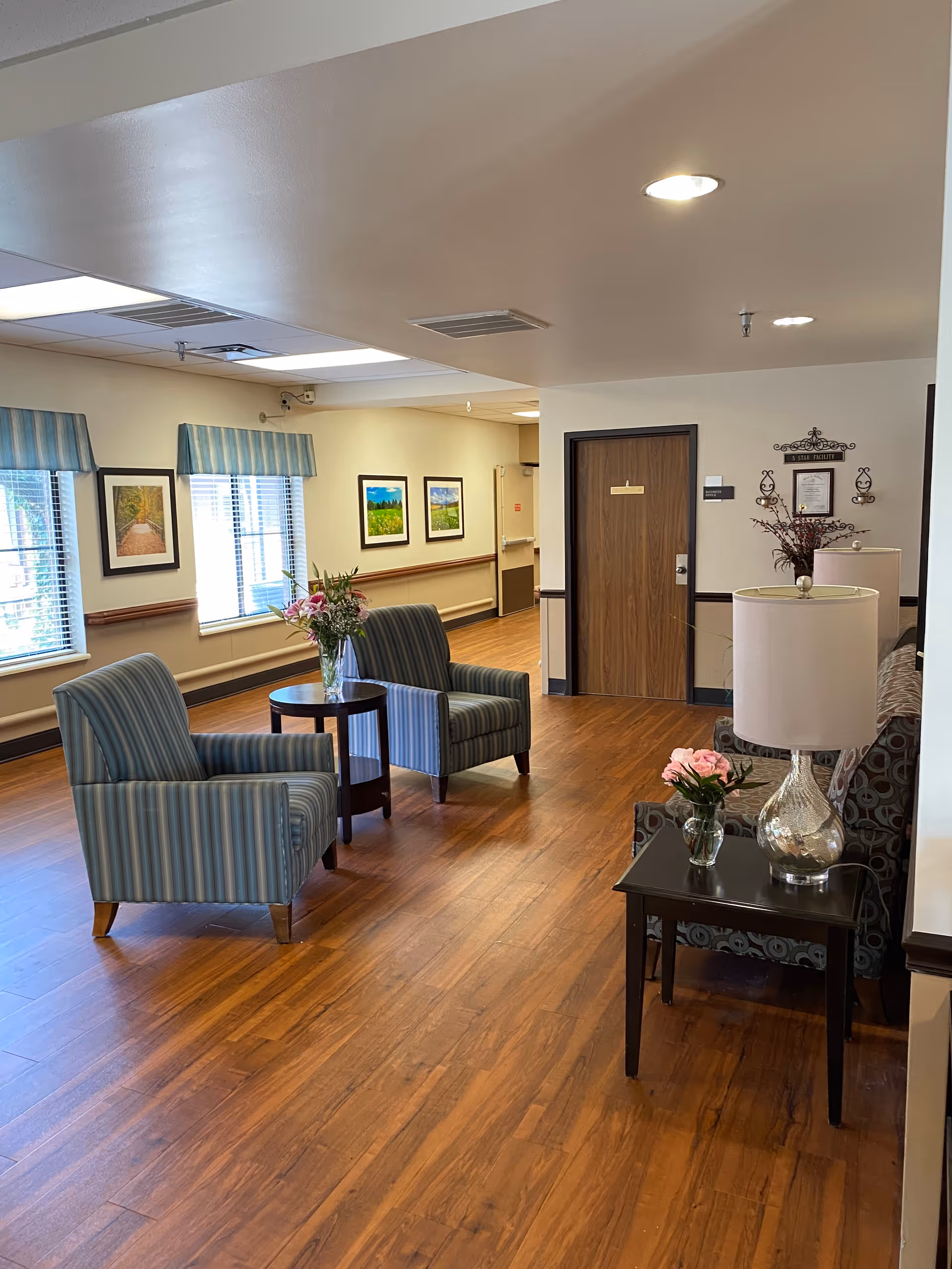 A cozy seating area in a senior living facility with two striped armchairs around a small round table holding a vase of flowers. Nearby is a side table with a lamp and a small vase of pink flowers. The room has wooden flooring, framed landscape pictures on the walls, and windows with striped valances letting in natural light.