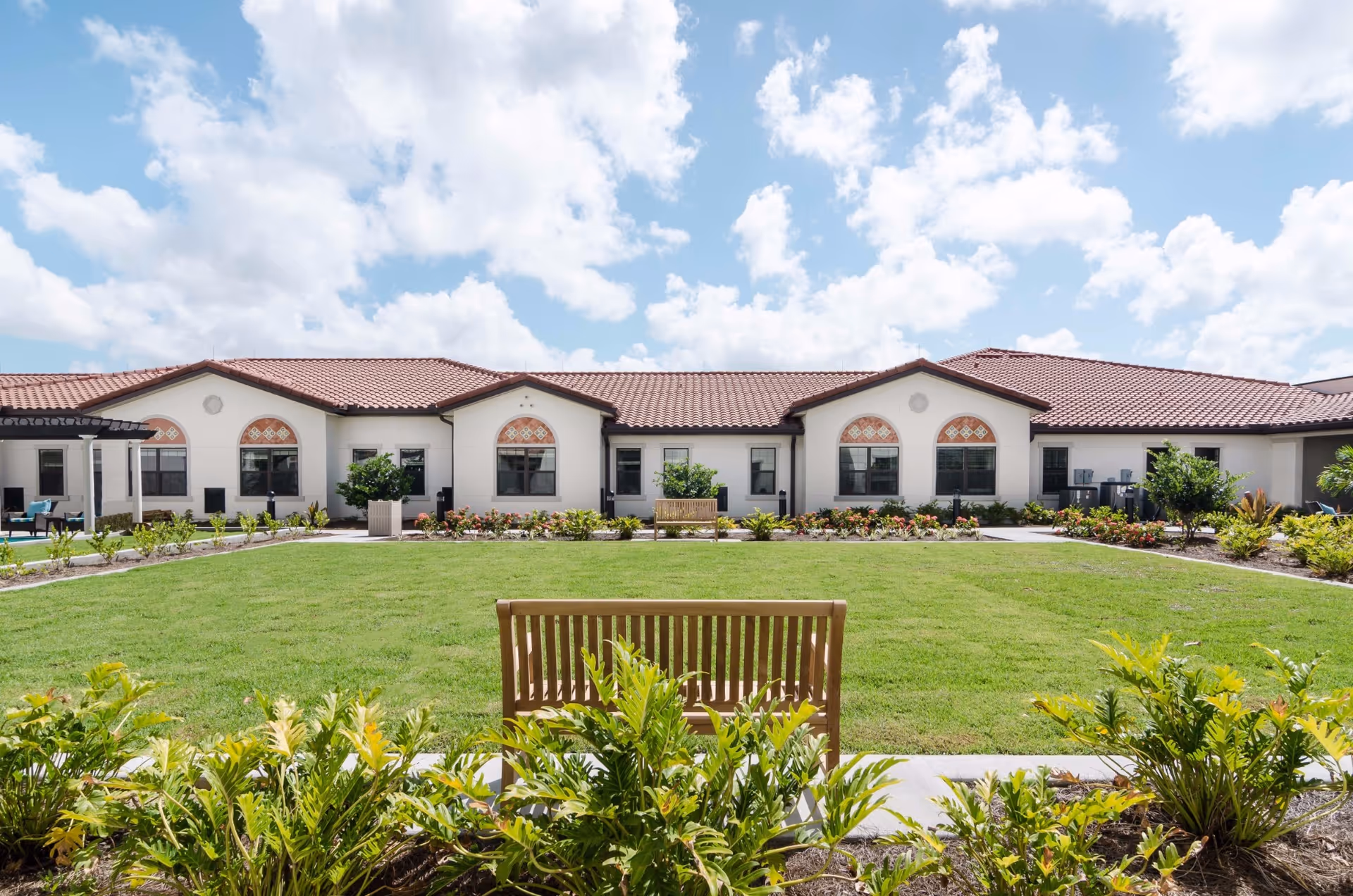 Outdoor garden area at American House Coconut Point featuring a well-maintained lawn, wooden benches, various plants and shrubs, and a building with a red tile roof and arched windows in the background under a partly cloudy sky.