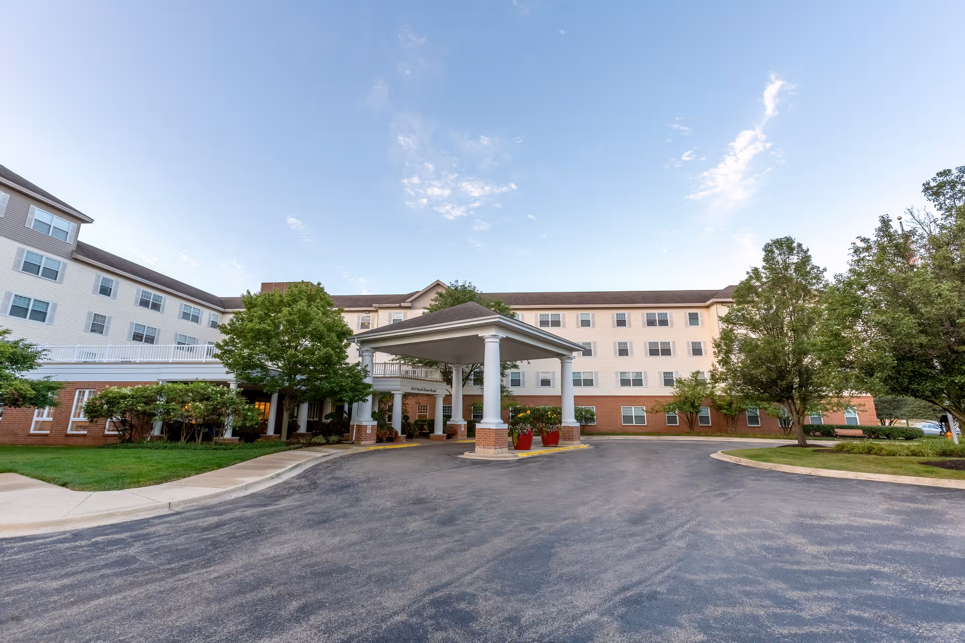 Exterior view of a multi-story senior living facility building with a covered entrance driveway, surrounded by trees and landscaping under a partly cloudy sky.
