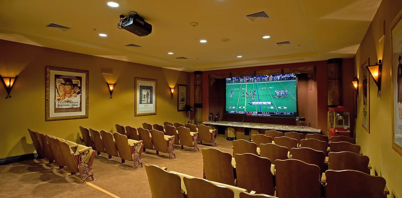 A small theater room with rows of wooden seats facing a large screen showing a football game. The room has warm lighting with wall sconces and framed movie posters on the walls. A popcorn machine is visible in the corner.