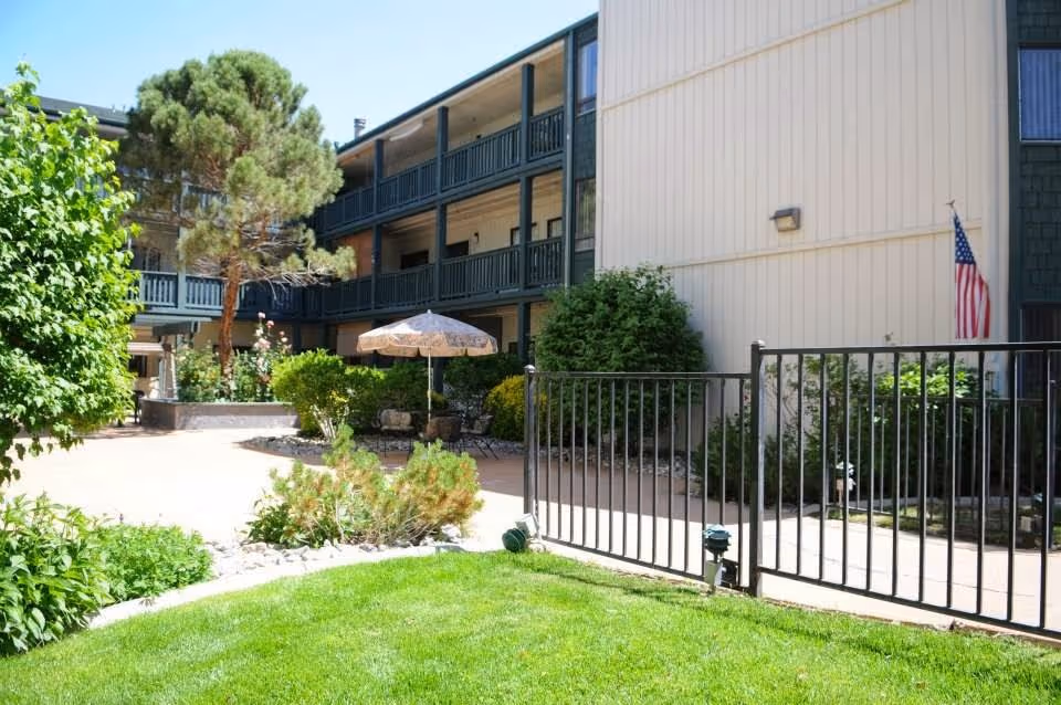 Outdoor courtyard area of a senior living facility with green grass, bushes, trees, a patio umbrella, and a multi-story building with balconies in the background. An American flag is visible on the right side near the building.