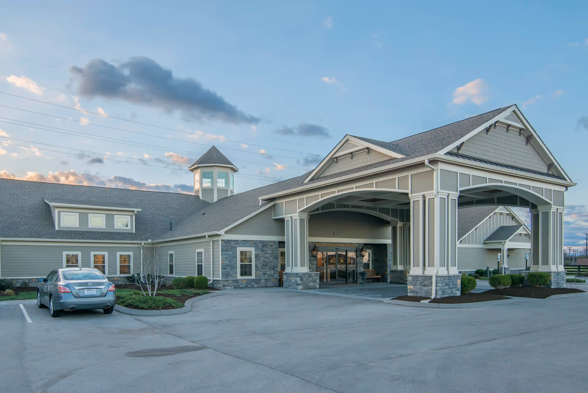 Exterior view of The Lantern at Morning Pointe Alzheimer’s Center of Excellence in Lexington showing a large covered entrance with columns, a stone and siding facade, a parked car, and a partly cloudy sky at dusk.