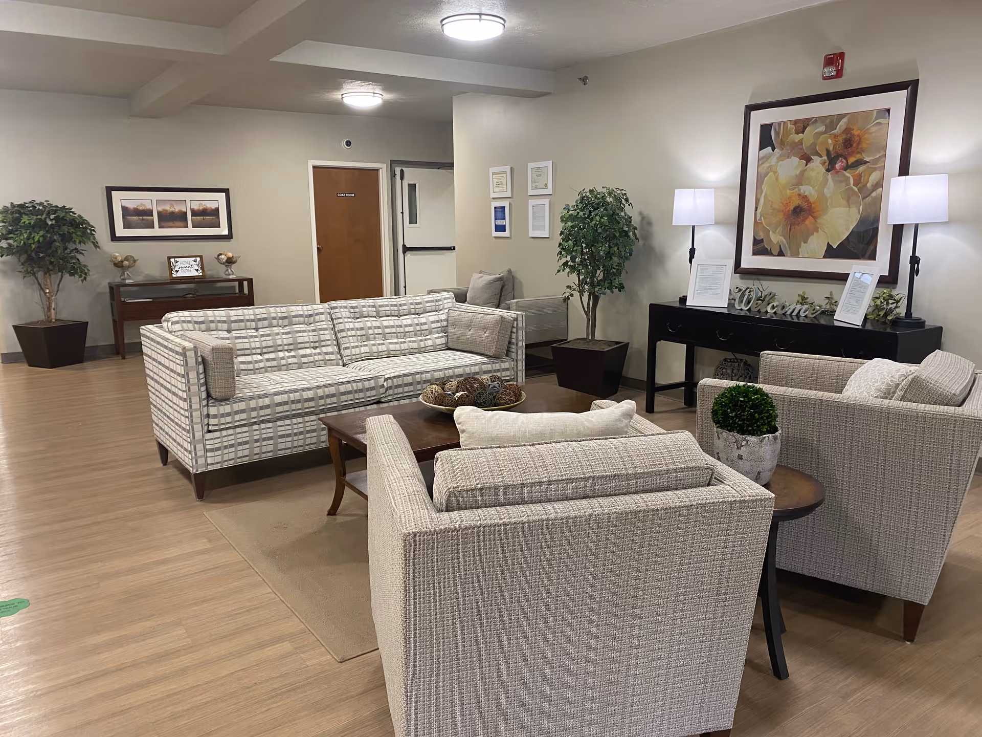 A cozy living room area in a senior living facility featuring a patterned sofa, two matching armchairs, a wooden coffee table with decorative items, and a side table with a small potted plant. The room has light-colored walls adorned with framed artwork and certificates, two floor lamps, and two large potted plants. The flooring is wood, and there is a door labeled 'Staff Room' in the background.