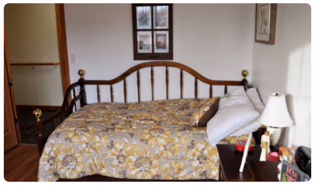 A bedroom with a wooden bed frame featuring brass finials, a floral patterned bedspread in shades of yellow and gray, white pillows, and a bedside table with a white lamp and various small items. There are framed pictures on the wall above the bed and a wooden door partially open to the left.