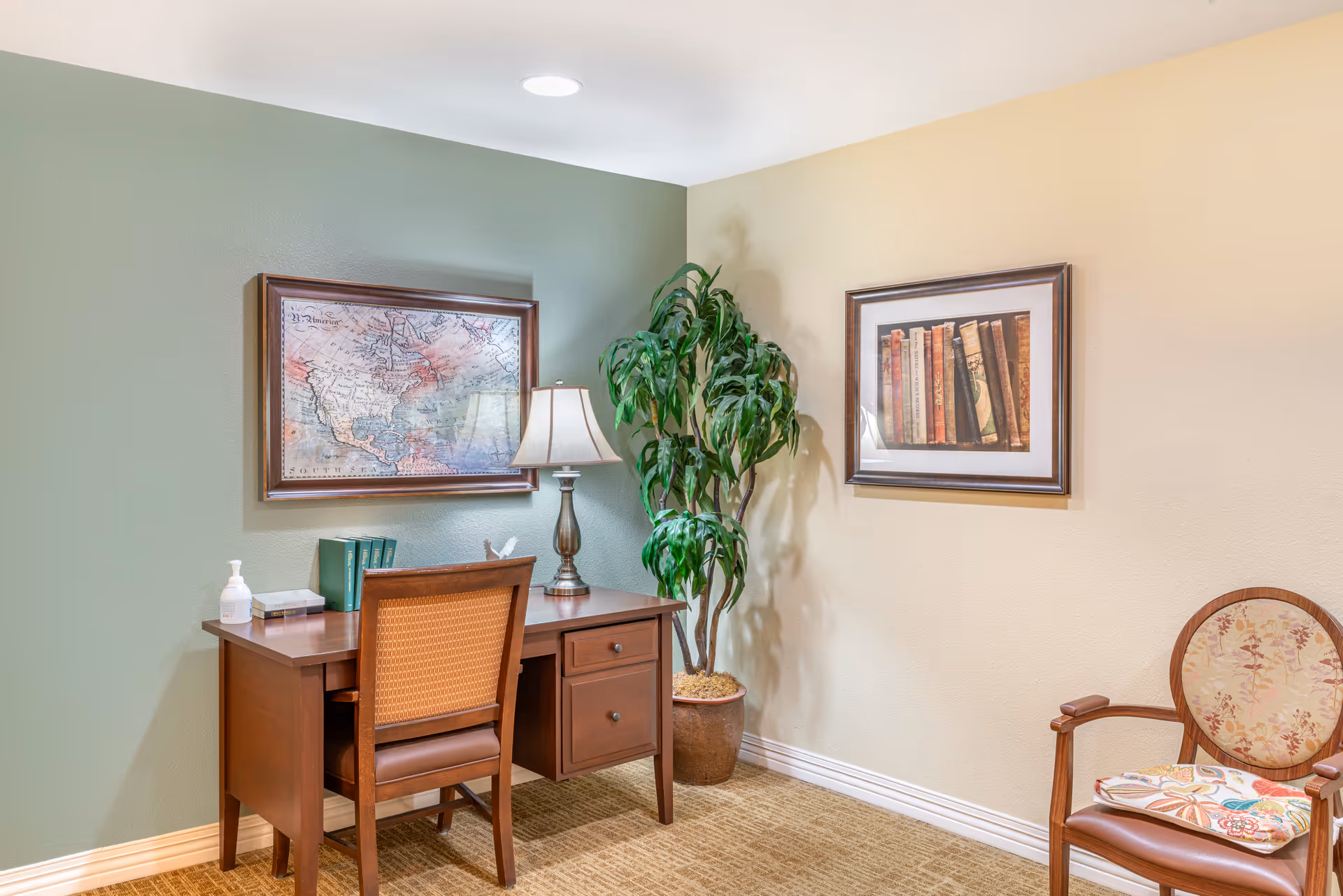 A cozy corner of a room featuring a wooden desk with a chair, a table lamp, and some books. On the wall above the desk is a framed vintage map. Next to the desk is a large potted plant, and on the adjacent wall is a framed picture of books. There is also an upholstered wooden chair with a floral cushion on the right side. The walls are painted in two tones: green and beige.